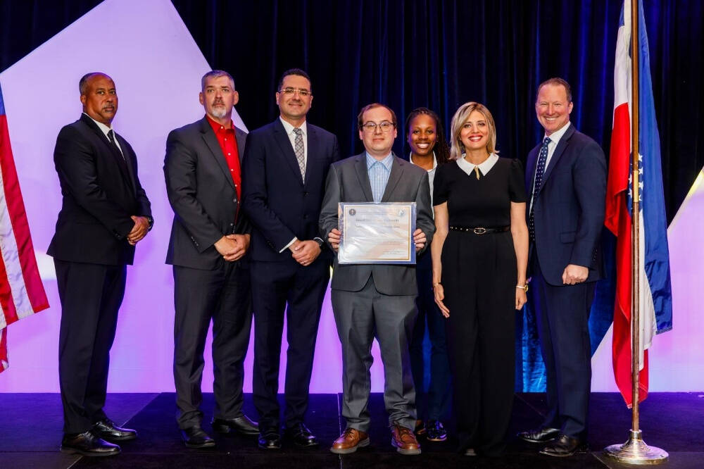 A group of seven people, dressed in business attire, stand on a stage between two flags. The person in the center is holding a framed certificate, and the group is posed in front of a dark curtain with geometric lighted shapes in the background.